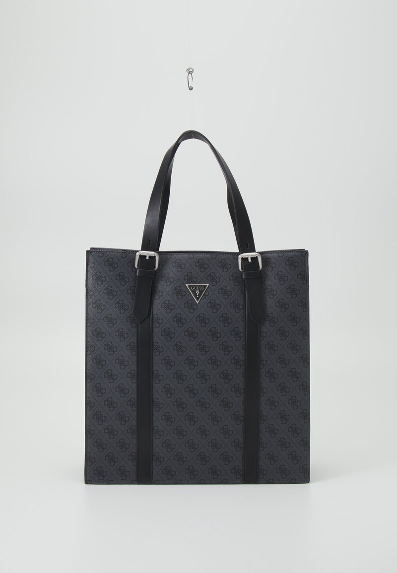 Schwarze Guess-Tasche mit Monogrammmuster, silbernen Schnallen und doppelten Schulterriemen, die vor einem schlichten weißen Hintergrund hängt.