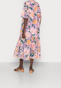 Vestido maxi floral en púrpura claro con grandes flores melocotón y amarillas, mangas cortas, dobladillo con volantes y un ajuste relajado. Llevado con sandalias en tono arena.