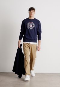 Młody mężczyzna spacerujący wewnątrz, ubrany w granatowy sweatshirt z logo, beżowe spodnie, białe sneakersy, trzymający czarną kurtkę w lewej ręce.