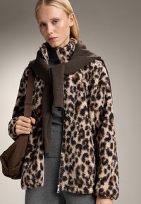 Giacca in pile con stampa leopardata in beige e marrone, caratterizzata da una chiusura con zip, vestibilità rilassata e tessuto testurizzato. Abbinata a un top grigio e pantaloni grigi.