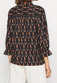 Blusa nera caratterizzata da un motivo ikat arancione e beige, maniche lunghe con polsini svasati e un dettaglio arricciato sul corpetto posteriore.