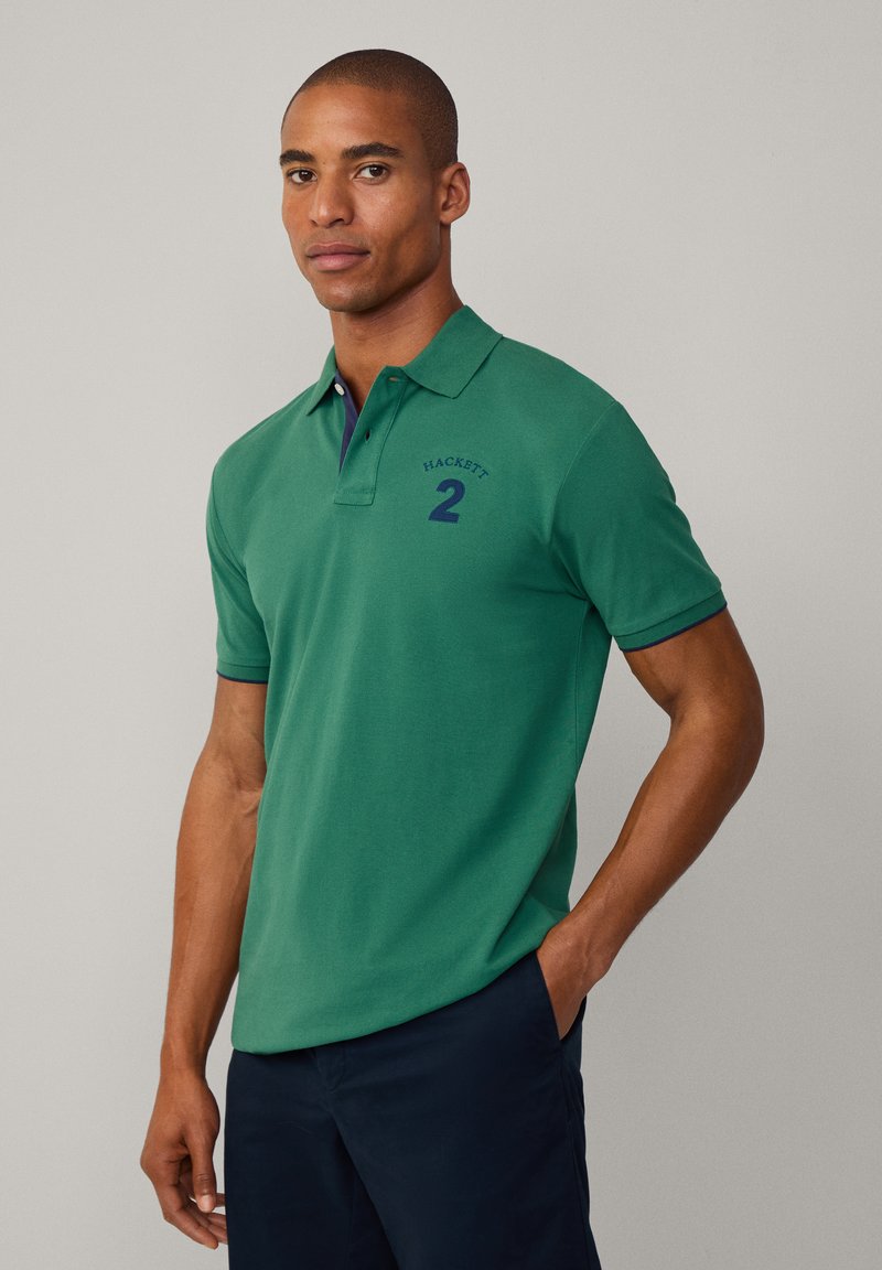 Grønn polo skjorte med krage, korte ermer, brodert "HACKETT" og nummer "2" i marineblå, bomullsstoff, glatt tekstur.