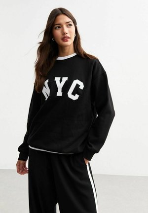 Czarna bluza z białym napisem "NYC", z prążkowanymi mankietami i okrągłym dekoltem, połączona z czarnymi spodniami z białymi paskami po bokach.
