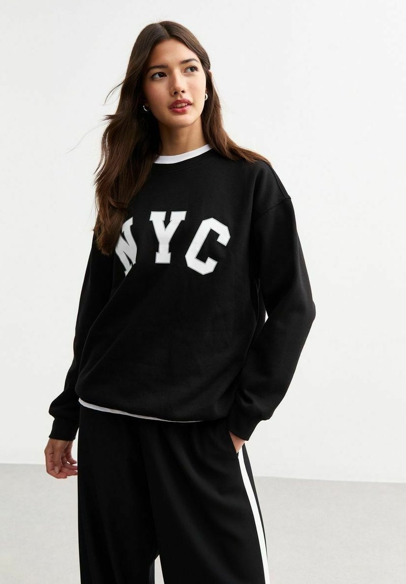 Sudadera negra con letras blancas "NYC", puños acanalados y cuello redondo, acompañada de pantalones negros con franjas laterales blancas.