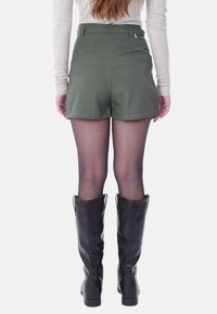 Shorts verde oliva a vita alta con tasche laterali, abbinati a stivali neri sopra il ginocchio e collant velati, il tessuto ha una consistenza liscia.