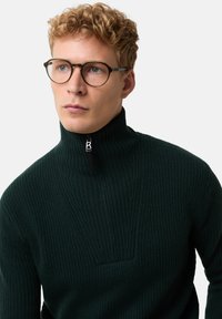 Junger Mann mit lockigem blondem Haar, trägt runde Brille und einen dunkelgrünen, gerippten Pullover mit einem Reißverschluss und hohem Kragen.