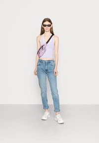 Top halter lilás claro, jeans azuis, ténis brancos e uma mochila multicolorida. Óculos de sol completam o visual. Fundo simples.