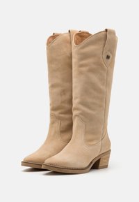 Botas de vaquero beige de ante hasta la rodilla con punta afilada, detalles de costura sutiles y tacón apilado. Presenta una lengüeta de agarre en la parte superior.