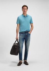 Himmelsblå polo-shirt, slidte blå jeans, brune læder loafers, og en sort duffelbag med top-håndtag; afslappet outfit vist mod en neutral baggrund.