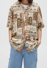 Personne portant une chemise à manches courtes boutonnée avec des motifs abstraits de feuilles et géométriques verts, marron et beige, et un pantalon en denim bleu clair.