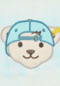 Diseño de cara de oso bordado en tela de color azul claro, con una gorra azul y un acento amarillo. Detalles suaves y texturizados en blanco en el oso.