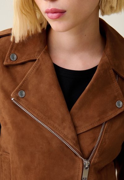 Giacca in suede marrone con dettagli in metallo argentato, caratterizzata da una zip asimmetrica e rever a punte, indossata sopra un top nero. Texture liscia.