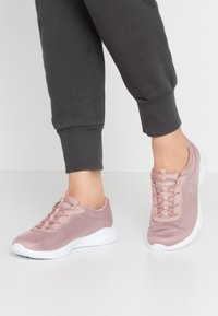 Světle růžové sportovní boty vyrobené z síťoviny, s designem na šněrování, bílou podrážkou a strukturovaným povrchem. Nošeny s šedými tepláky.