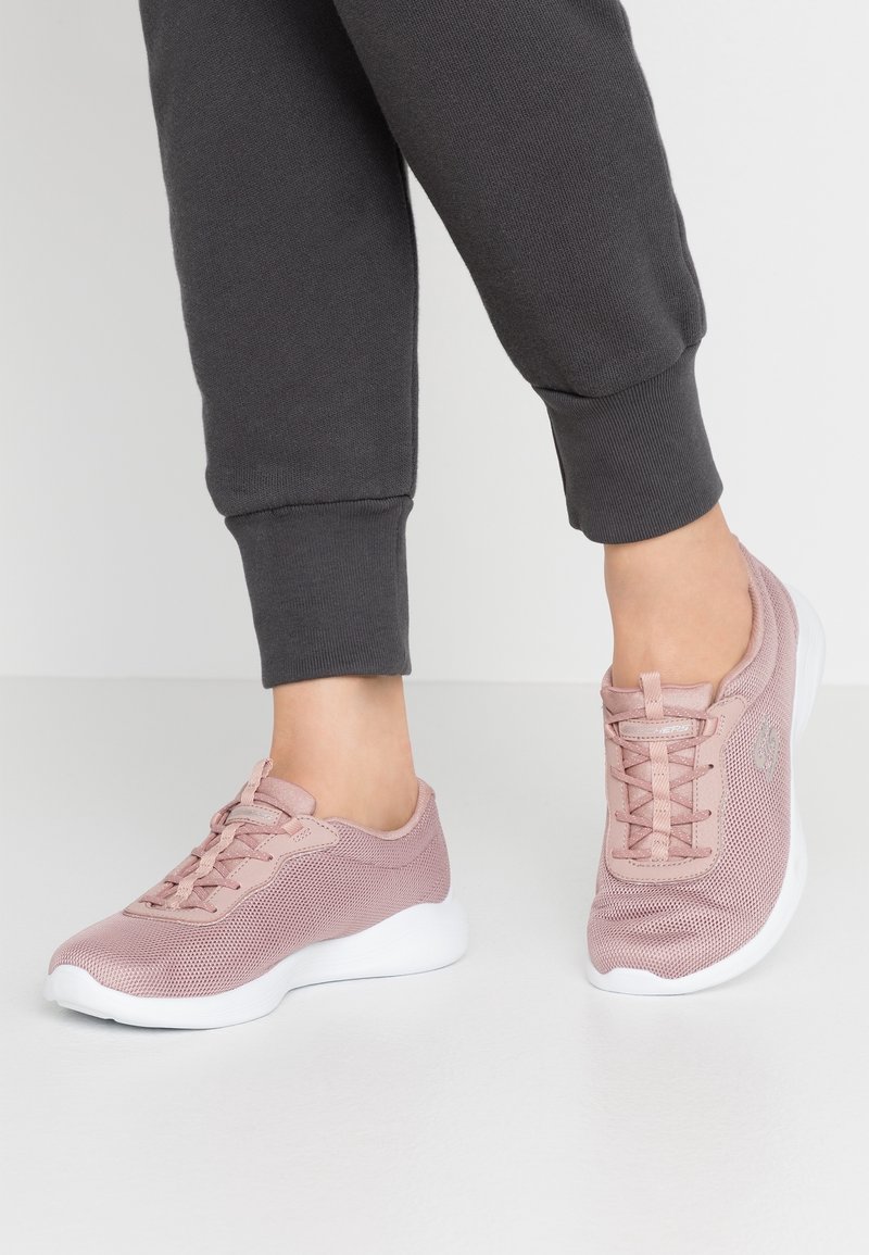 Světle růžové sportovní boty vyrobené z síťoviny, s designem na šněrování, bílou podrážkou a strukturovaným povrchem. Nošeny s šedými tepláky.