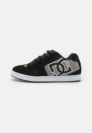 Black sneakers with a textured gray panel, featuring a white logo. Made of suede and fabric, they have a white rubber sole and black laces.