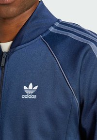 Navy blue zip-up jacket made of smooth fabric. Features white Adidas logo and three light blue stripes along the shoulders.