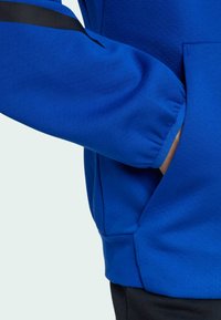 Blå sweatshirt med kenguruficka, elastiska ärmslut och en svart accentrand på ärmen. Tillverkad av mjukt, strukturerat tyg.