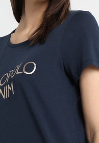 Námořnicky modré tričko z bavlny s krátkými rukávy, s metalickým zlatým potiskem, na kterém je moderním písmem napsáno "TOP OLO".