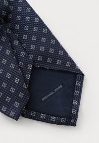 Marineblau gemusterte Krawatte aus strukturiertem Stoff, mit einem geometrischen Design in Weiß und Grau. Auf der Unterseite steht das Label "Michael Kors".