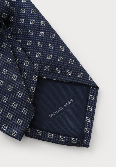 Cravate à motif bleu marine en tissu texturé, arborant un design géométrique en blanc et gris. L'étiquette indique "Michael Kors" sur le dessous.
