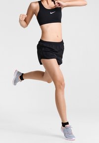 Svart sport-bh och svarta träningsshorts, kombinerat med ljusgrå träningssneakers med rosa detaljer, mot en neutral bakgrund.