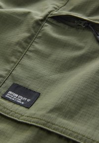 Tessuto verde oliva con una trama a griglia, con un'etichetta del marchio nera cucita. Include una tasca con cerniera lungo la cucitura.