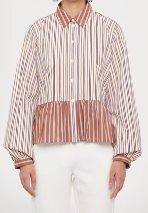 Vrouw die een blouse met lange mouwen en knopen draagt, met verticale strepen in wit, roestkleur, geel en paars, gecombineerd met een witte broek.