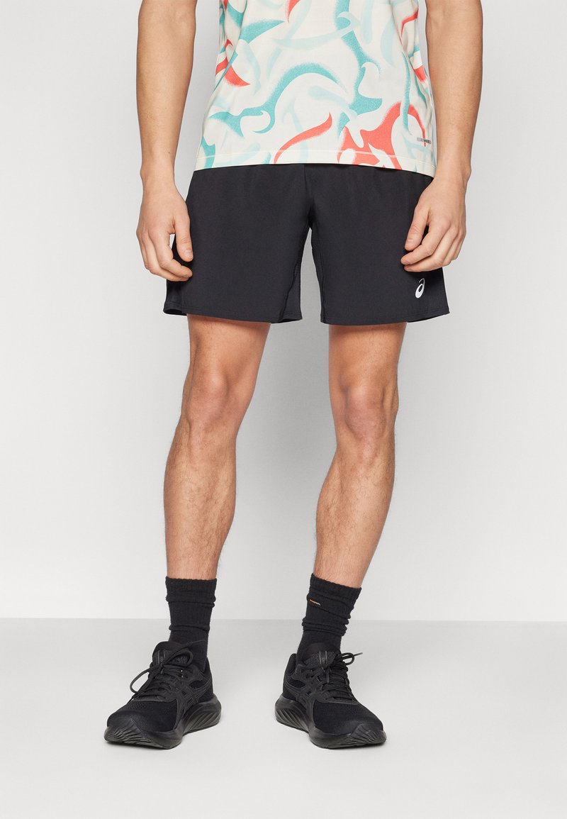 Zwarte sportshorts met een gladde textuur, gecombineerd met een kleurrijke, patroondruppel-shirt. Zwarte sneakers en sokken maken het outfit compleet.