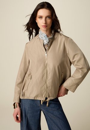 Mujer de cabello oscuro con una chaqueta beige con cremallera, jeans azules, una bufanda con estampado y una pulsera dorada, de pie frente a un fondo neutro.