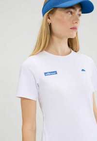 T-shirt bianco a maniche corte con accenti di logo blu. Tessuto morbido, scollatura rotonda, dettagli sulle cuciture laterali. Il modello indossa un berretto blu.