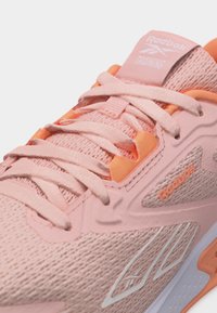 Nærbillede af pink og orange Reebok træningssneaker med struktureret stof, snørebånd og mærkelogoer på tunge og sidepanel.