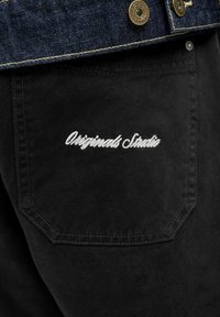 Gros plan sur la poche d’un pantalon en denim noir avec une broderie blanche « Originals Studio » sous une veste en denim bleu foncé avec des boutons en laiton.