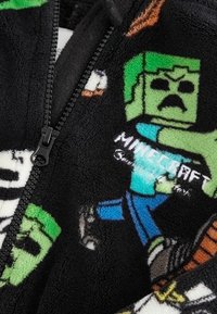 Giacca in pile nera con zip, caratterizzata da stampe colorate di personaggi di Minecraft. Dettagli notevoli includono un tessuto testurizzato e una zip nera.