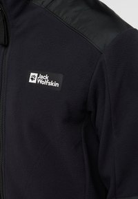 Giacca in pile nera con pannelli sulle spalle testurizzati; presenta una toppa con il logo bianco di Jack Wolfskin sul petto. Chiusura con zip.