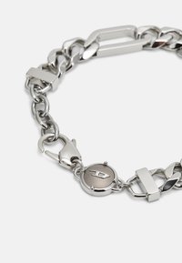 Silvermetall armband med kedjor som har sammanlänkade ovala och rektangulära former, en central logodisks och en säker låsmekanism.