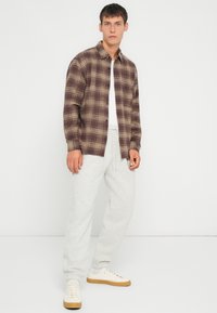 Camisa de franela a cuadros marrón y beige sobre una camiseta blanca, combinada con pantalones de chándal gris claro y zapatillas color crema. Estilo básico, ajuste relajado.
