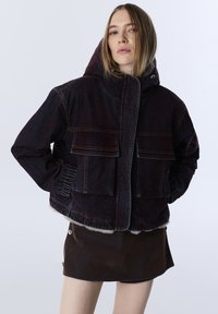 Tmavě modrá džínová bunda s kapucí, vybavená dvěma našitými kapsami, ribbed manžetami a měkkým kožíškem na spodní části pro přidanou texturu.