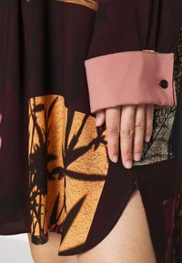 Main avec des ongles naturels reposant sur l'ourlet d'une robe sombre à motifs avec large poignet rose et détail bouton sur la manche.