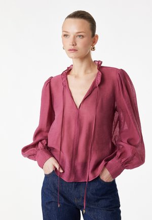 Donna con una blusa trasparente magenta con colletto arricciato e maniche lunghe a sbuffo, abbinata a jeans blu scuro, con le mani nelle tasche.