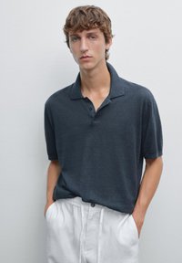 Czarna szara koszulka polo z krótkim rękawem, cechująca się luźnym krojem, zapięciem na dwa guziki oraz miękką, teksturowaną tkaniną. W zestawie z białymi lnianymi spodniami.