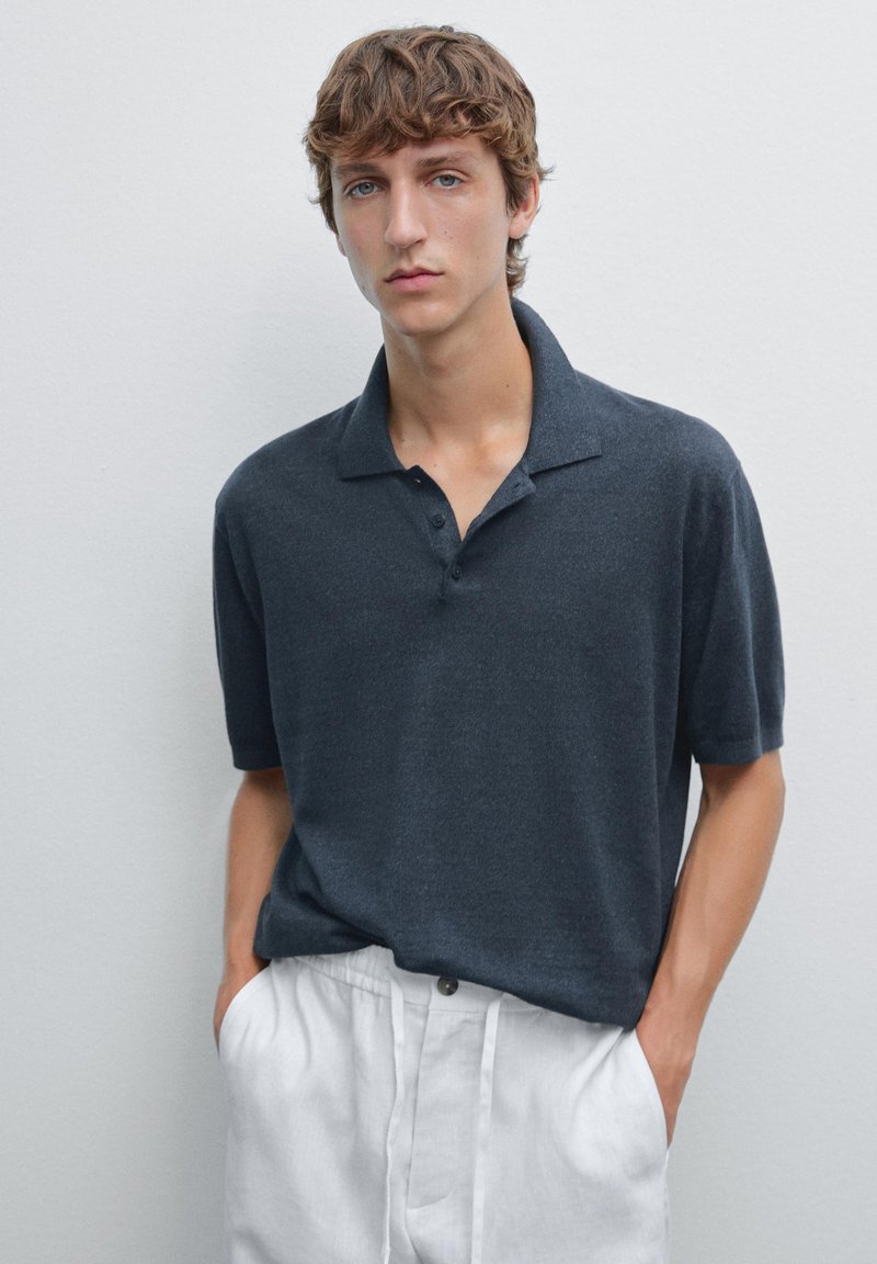 Czarna szara koszulka polo z krótkim rękawem, cechująca się luźnym krojem, zapięciem na dwa guziki oraz miękką, teksturowaną tkaniną. W zestawie z białymi lnianymi spodniami.