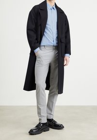 Manteau oversized noir, chemise boutonnée bleu clair, pantalon ajusté gris et mocassins noirs brillants avec des semelles épaisses. Textures lisses, coupe ajustée.