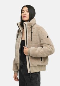 Beige gewatteerde jas met een textuur van fleece, staande kraag, ritssluiting aan de voorkant en zwarte accenten op de zakken en trekkoorden.