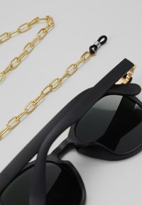 Black sunglasses with green lenses, featuring a chunky design. Accompanied by a gold chain link strap and a black rubber connector.