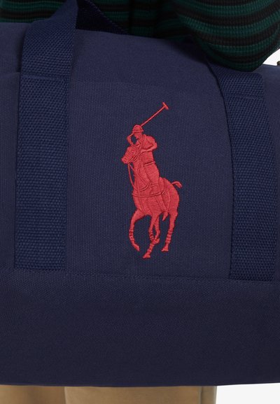 Marineblå Duffelbag med teksturert stoff, med en stor rød polo-spillerlogo brodert på siden. Robuste stropper, kompakt design.