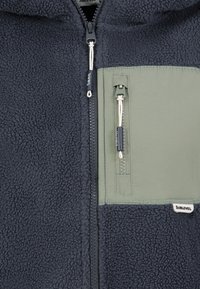 Gros plan d'une veste polaire gris foncé avec une fermeture éclair sur le devant et une poche poitrine zippée verte avec cordons.