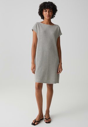 Frau mit lockigem Haar, die ein knielanges, schwarz-weiß gestreiftes Kleid mit kurzen Ärmeln und schwarzen Sandalen trägt, steht vor einem einfarbigen Hintergrund.