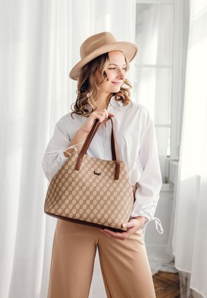 Beige patterned handbag with dark brown handles and logo, paired with a white blouse and light-colored trousers, set against a bright background.