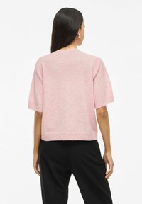 Maglione rosa chiaro a maniche corte con scollo rotondo e design corto. Texture morbida con un sottile motivo melangiato; indossato con pantaloni neri.