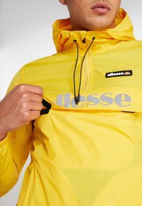 Personne portant une veste jaune à capuche, tirant une patte de fermeture éclair à l'avant révélant le nom de la marque ellesse en impression réfléchissante.